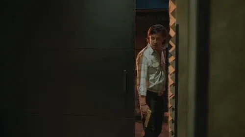Homem de camisa clara e calça escura,segurando uma pistola dourada,espreita numa abertura escura com olhar tenso.