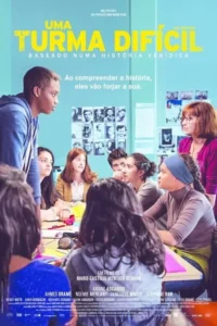Jovem em pé e professora observam alunos diversos sentados em mesa de aula,com fotos na parede. Clima de aprendizado e debate.