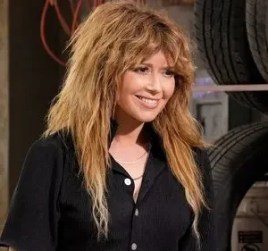 Mulher sorridente com cabelo em camadas bagunçado, vestindo camisa escura, em uma oficina com pneus empilhados ao fundo.