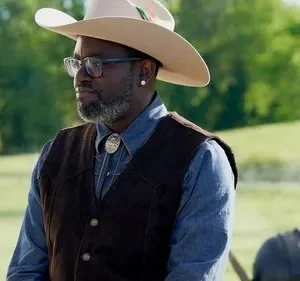 Homem negro de chapéu cowboy claro, óculos e colete marrom, com barba grisalha, em campo verde ensolarado.