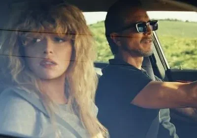 Mulher loira encaracolada e homem barbudo de óculos dirigem. Ela está pensativa, ele sorri levemente, sob sol em paisagem rural.