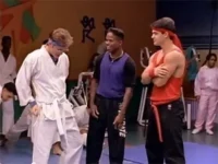 Três jovens num dojo. Um de gi branco, um de roxo e um de vermelho com faixa, confiante. Outras pessoas treinam ao fundo.