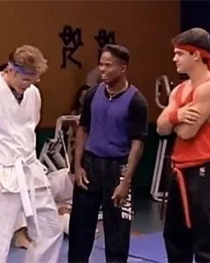 Três jovens num dojo. Um de gi branco, um de roxo e um de vermelho com faixa, confiante. Outras pessoas treinam ao fundo.