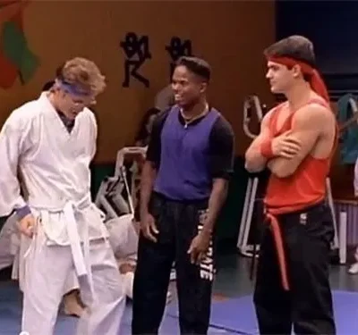 Três jovens num dojo. Um de gi branco, um de roxo e um de vermelho com faixa, confiante. Outras pessoas treinam ao fundo.