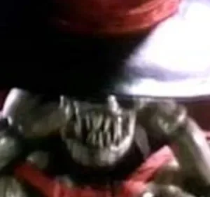 Criatura bizarra de pele escura e dentes afiados, carregando um objeto metálico gigante, em ambiente sombrio com tons vermelhos.