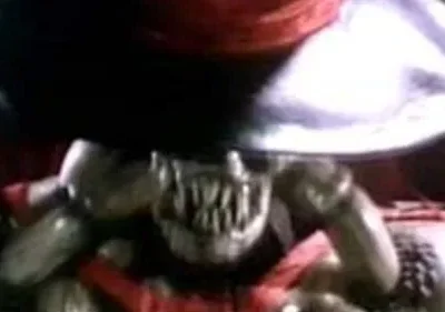 Criatura bizarra de pele escura e dentes afiados, carregando um objeto metálico gigante, em ambiente sombrio com tons vermelhos.