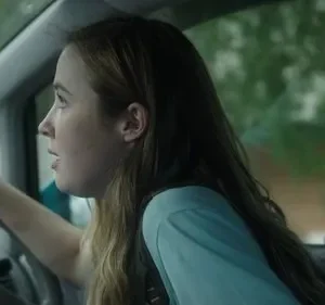 Uma jovem com cabelo castanho claro dirige um carro, com a boca ligeiramente aberta e expressão de surpresa ou preocupação. O exterior verde é borrado.