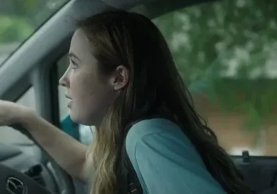 Uma jovem com cabelo castanho claro dirige um carro, com a boca ligeiramente aberta e expressão de surpresa ou preocupação. O exterior verde é borrado.