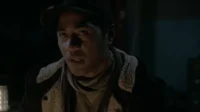 Homem de boné e jaqueta, com o rosto iluminado na escuridão. Sua expressão é intensa, boca ligeiramente aberta, como se falasse.