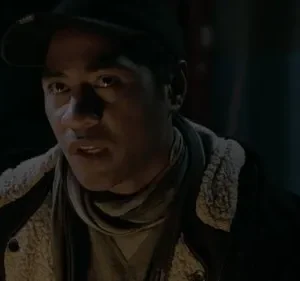 Homem de boné e jaqueta, com o rosto iluminado na escuridão. Sua expressão é intensa, boca ligeiramente aberta, como se falasse.