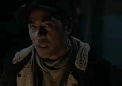 Homem de boné e jaqueta, com o rosto iluminado na escuridão. Sua expressão é intensa, boca ligeiramente aberta, como se falasse.