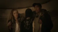 Três pessoas em um túnel escuro: mulher preocupada, homem apoiando alguém com camiseta de caveira. Ambiente sombrio, clima tenso.