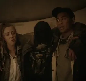 Três pessoas em um túnel escuro: mulher preocupada, homem apoiando alguém com camiseta de caveira. Ambiente sombrio, clima tenso.