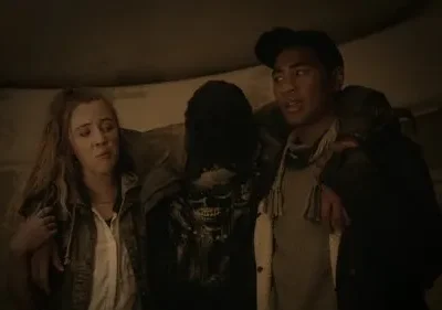 Três pessoas em um túnel escuro: mulher preocupada, homem apoiando alguém com camiseta de caveira. Ambiente sombrio, clima tenso.