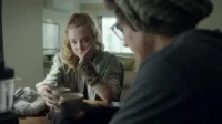 Mulher loira olha pensativa para frente, segurando uma caneca, enquanto um homem de óculos e gorro está de costas. Clima sério.