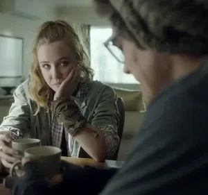 Mulher loira olha pensativa para frente, segurando uma caneca, enquanto um homem de óculos e gorro está de costas. Clima sério.