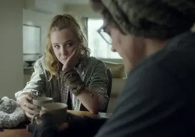 Mulher loira olha pensativa para frente, segurando uma caneca, enquanto um homem de óculos e gorro está de costas. Clima sério.