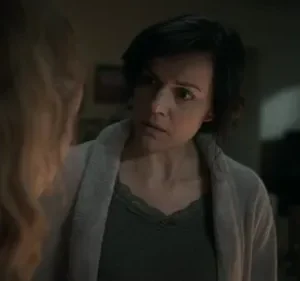 Mulher de cabelo escuro com expressão séria olha para outra de costas. Ambiente doméstico escuro, clima de preocupação.