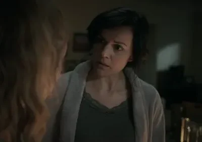 Mulher de cabelo escuro com expressão séria olha para outra de costas. Ambiente doméstico escuro, clima de preocupação.