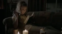 Jovem com cabelo claro lê um livro em um sofá, iluminada por lanterna e velas. Ambiente escuro e misterioso.