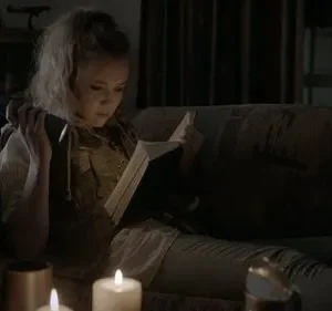 Jovem com cabelo claro lê um livro em um sofá, iluminada por lanterna e velas. Ambiente escuro e misterioso.