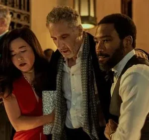 Mulher de vestido vermelho com olhos fechados é amparada por dois homens de terno em festa elegante e tensa.