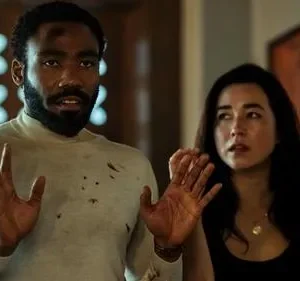 Um homem barbudo com camisa rasgada e mulher assustada, ambos com mãos levantadas, num ambiente interno tenso.