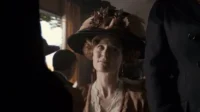 Mulher de chapéu elaborado e traje de época, com olhar pensativo. Cena interior histórica com luz dramática e outras figuras.