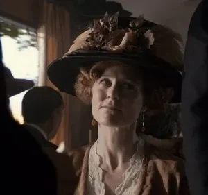 Mulher de chapéu elaborado e traje de época, com olhar pensativo. Cena interior histórica com luz dramática e outras figuras.