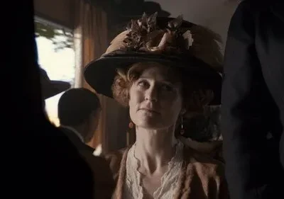 Mulher de chapéu elaborado e traje de época, com olhar pensativo. Cena interior histórica com luz dramática e outras figuras.