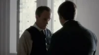 Dois homens em trajes de época. Um de costas, outro sério com colete e camisa listrada, em uma sala com luz difusa. Clima sóbrio.
