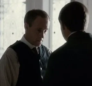 Dois homens em trajes de época. Um de costas, outro sério com colete e camisa listrada, em uma sala com luz difusa. Clima sóbrio.