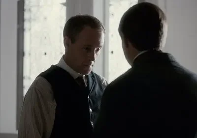 Dois homens em trajes de época. Um de costas, outro sério com colete e camisa listrada, em uma sala com luz difusa. Clima sóbrio.