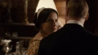 Mulher em vestido dourado e faixa na cabeça sorri para homem de terno. Cenário interior sofisticado com pouca luz.
