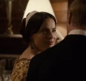 Mulher em vestido dourado e faixa na cabeça sorri para homem de terno. Cenário interior sofisticado com pouca luz.