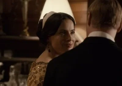 Mulher em vestido dourado e faixa na cabeça sorri para homem de terno. Cenário interior sofisticado com pouca luz.