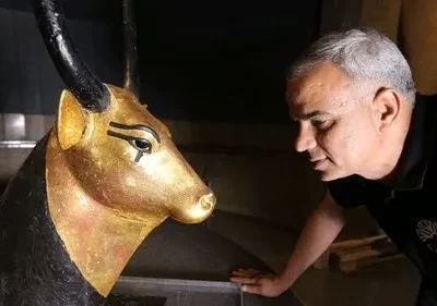 Homem com camisa preta examina um busto dourado de touro com chifres pretos e olhos egípcios. Fundo escuro, clima de descoberta.