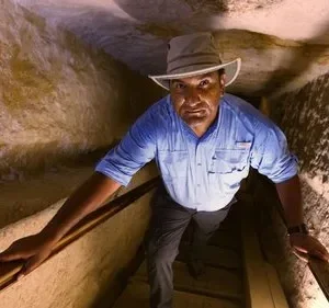 Um explorador de chapéu e camisa azul sobe um túnel rochoso e estreito, iluminado, com expressão intensa.