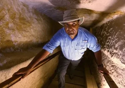 Um explorador de chapéu e camisa azul sobe um túnel rochoso e estreito, iluminado, com expressão intensa.