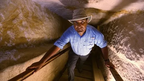 Um explorador de chapéu e camisa azul sobe um túnel rochoso e estreito,iluminado,com expressão intensa.