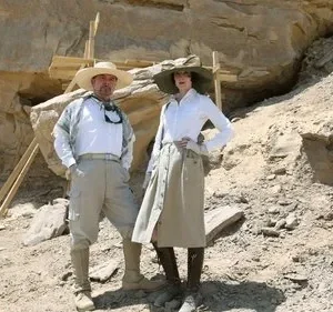 Um homem e uma mulher, vestidos como exploradores, posam em um sítio de escavação rochoso no deserto. Atmosfera de aventura.