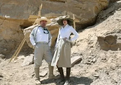 Um homem e uma mulher, vestidos como exploradores, posam em um sítio de escavação rochoso no deserto. Atmosfera de aventura.