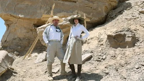 Vale dos Reis:Tesouros do Egito – T01E03:A Tumba Perdida de Cleópatra 1 Um homem e uma mulher,vestidos como exploradores,posam em um sítio de escavação rochoso no deserto. Atmosfera de aventura.