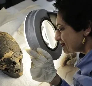 Uma cientista com luvas examina uma cabeça mumificada com uma lupa. Clima de mistério, pesquisa arqueológica e descobertas antigas.