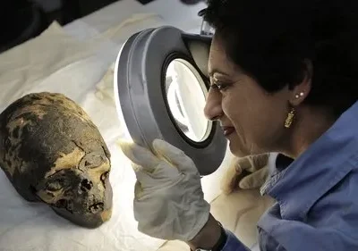 Uma cientista com luvas examina uma cabeça mumificada com uma lupa. Clima de mistério, pesquisa arqueológica e descobertas antigas.