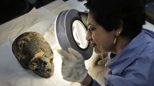 Vale dos Reis:Tesouros do Egito – T01E04:As Últimas Tumbas 1 Uma cientista com luvas examina uma cabeça mumificada com uma lupa. Clima de mistério,pesquisa arqueológica e descobertas antigas.