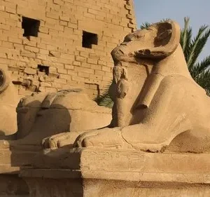 Estátuas monumentais de esfinges com cabeça de carneiro em fila, diante de ruínas de templo sob sol forte.