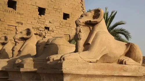 Estátuas monumentais de esfinges com cabeça de carneiro em fila,diante de ruínas de templo sob sol forte.
