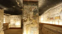 Tumba egípcia. Pilar e paredes com murais coloridos, figura divina central. Luz quente. Clima misterioso.