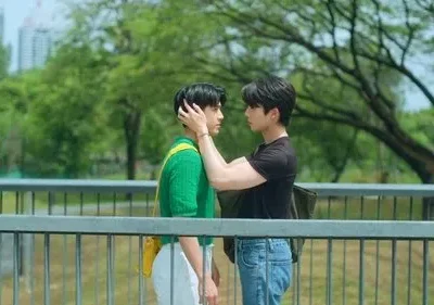 Dois jovens em momento terno numa passarela. Um toca o rosto do outro, com árvores e prédios ao fundo.